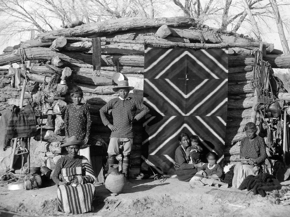 Black-and-white photo of a Navajo family in front of a log hogan with a woven blanket hanging by the door