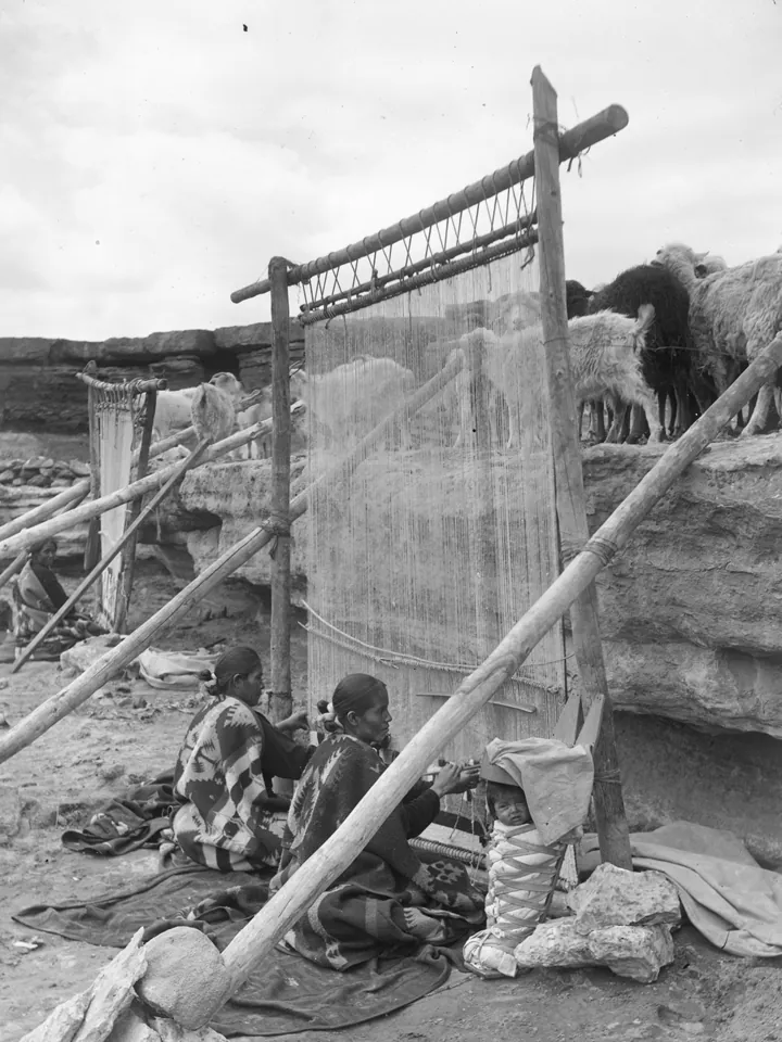 Black-and-white photo of Diné women beside a tall upright loom outdoors