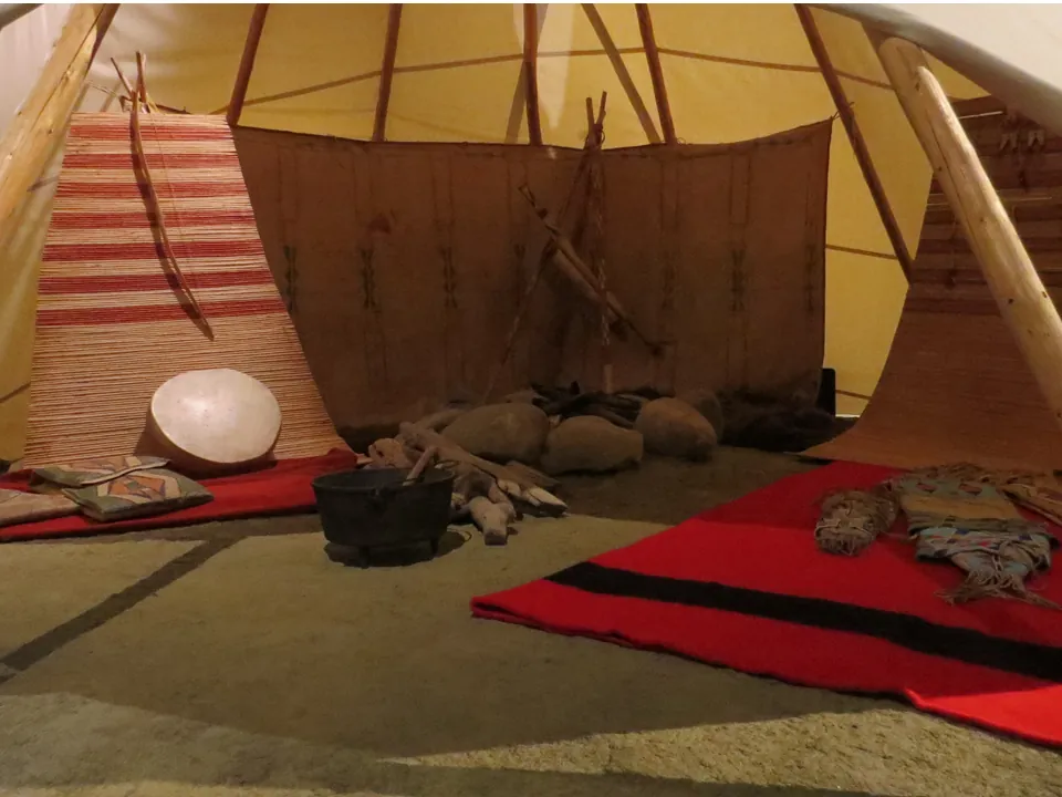Inside a tipi in a museum, with backrests, robes, and a red blanket