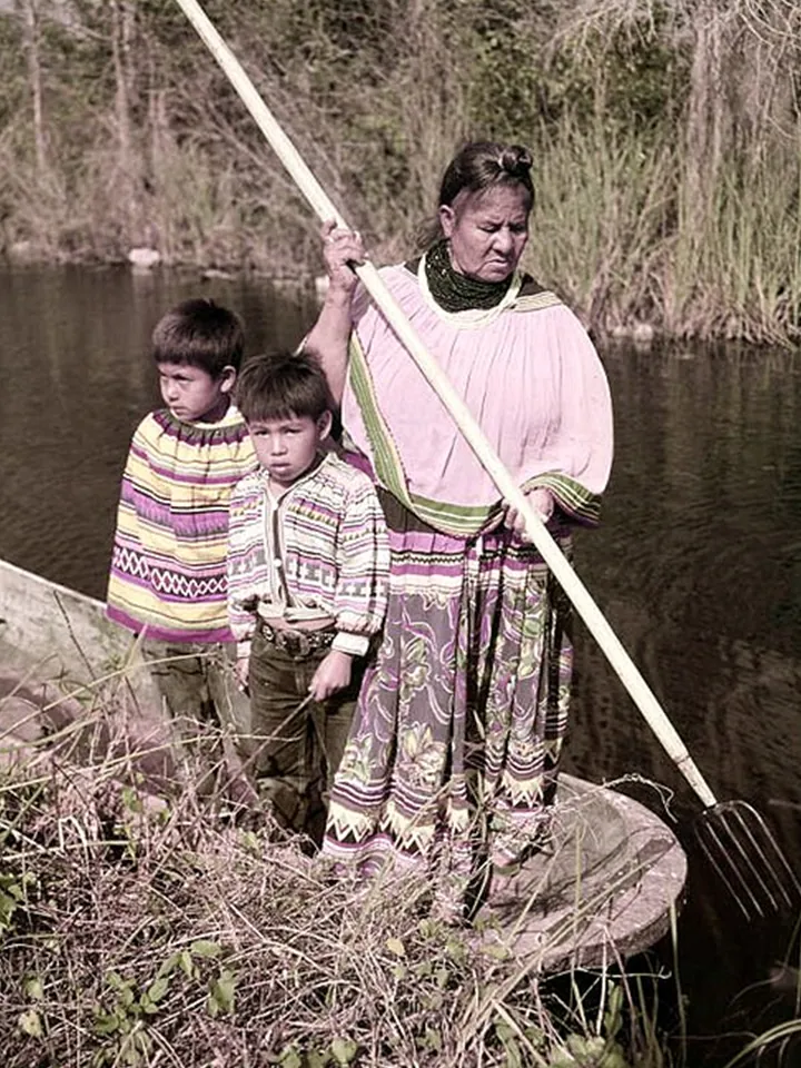 A Seminole woman and two boys standing in a dugout canoe; the woman holds a long pole with a pronged gig